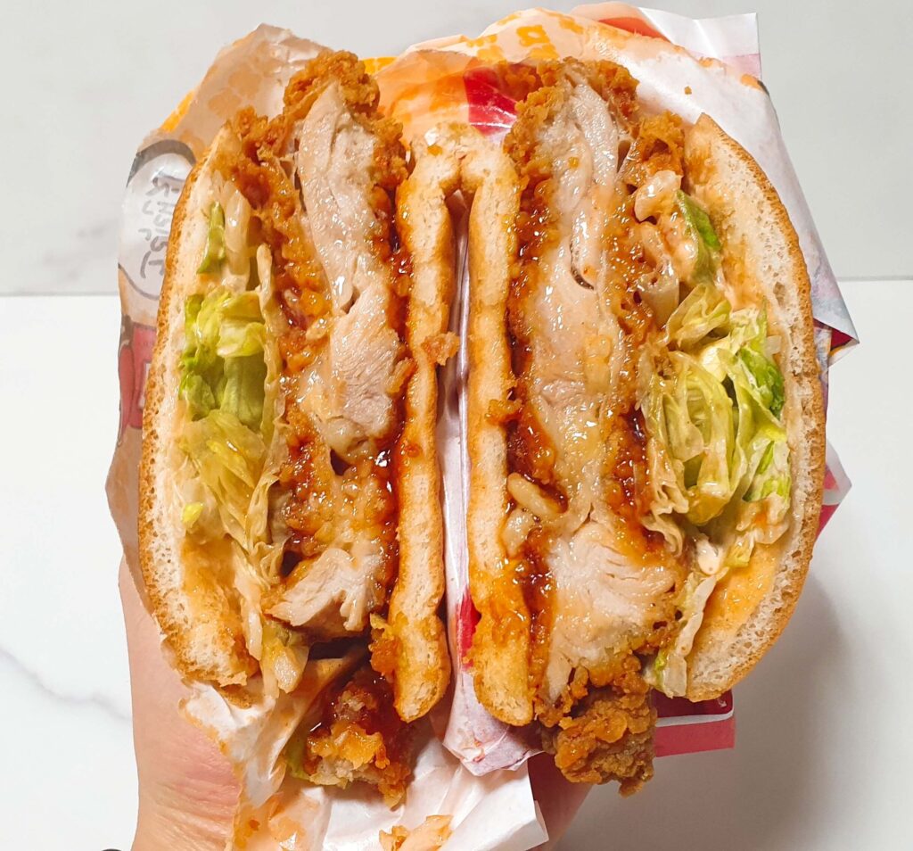 롯데리아 신메뉴 통다리 크리스피치킨버거 파이어핫을 반으로 잘라 두툼한 치킨 패티와 매콤한 레드 소스, 양상추의 조화를 보여주는 단면 사진